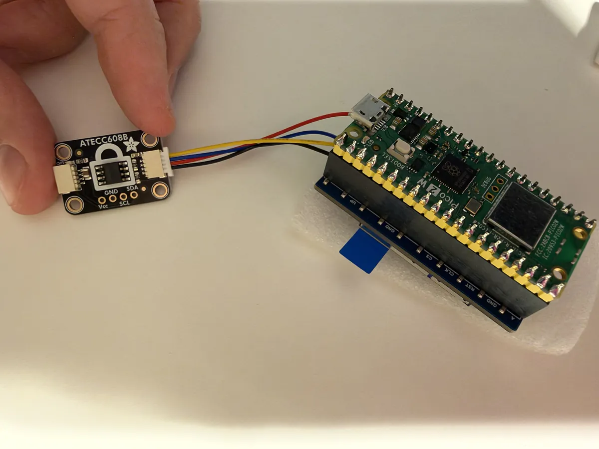 A hand holding the small chip board with the white plug pushed in, wired to the Pico